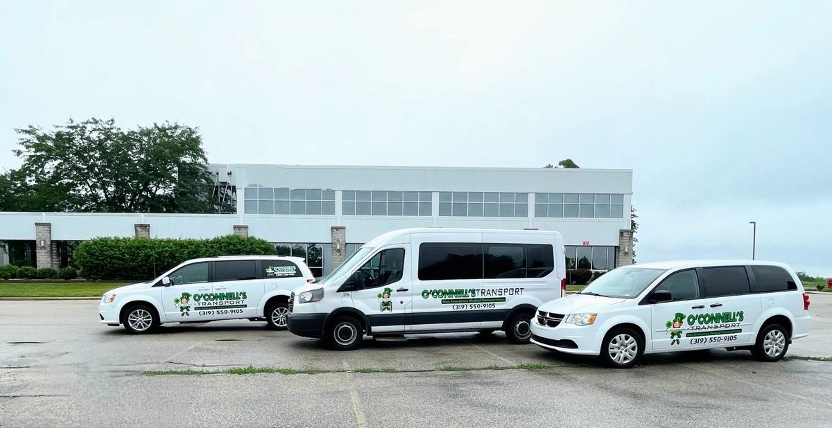 O'Connell's Transport's white vans with the green leprechaun logo, lined up in a parking lot: rear-entry wheelchair-accessible minivans and a larger passenger van